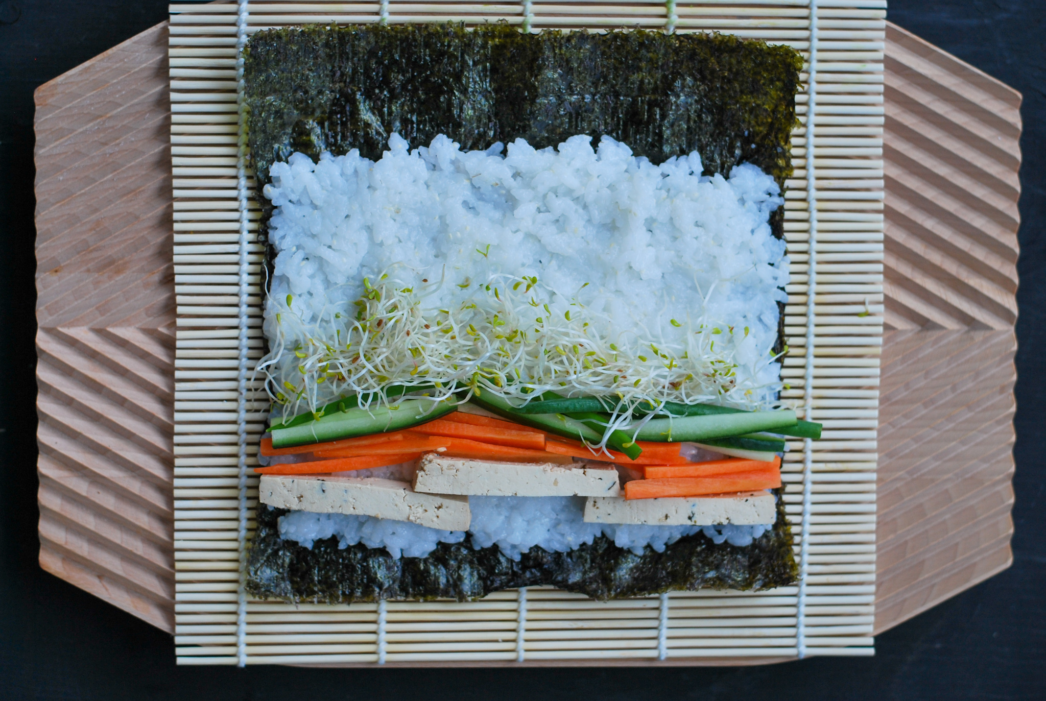Kuvassa on bambumaton päällä rullaamaton makirulla, jossa on riisiä, tofua, porkkanaa, kurkkua ja sinimailasen ituja.