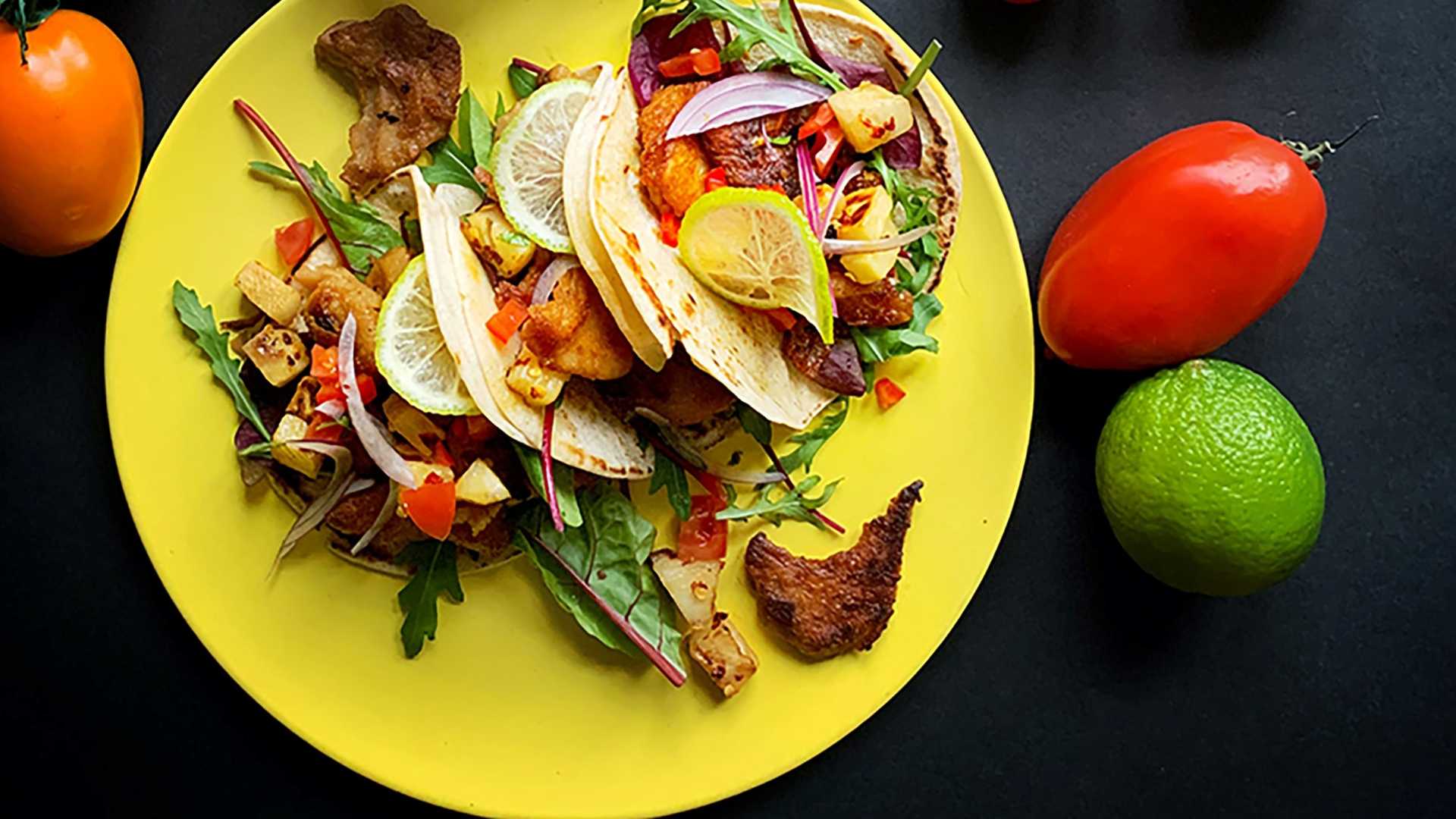 Kuvassa on keltainen lautanen, jonka päällä on kasviksilla ja kasviproteiinilla täytettyjä pieniä, rullaamattomia tortillalettuja. Lautasen vieressä on punainen ja oranssi tomaatti sekä lime.