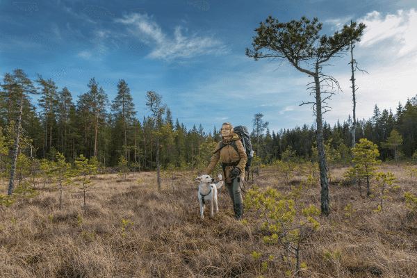 Kuvassa on Mia koiransa kanssa. Taustalla näkyy metsän raja sekä sininen taivas, jossa on pieniä pilvenrepaleita.