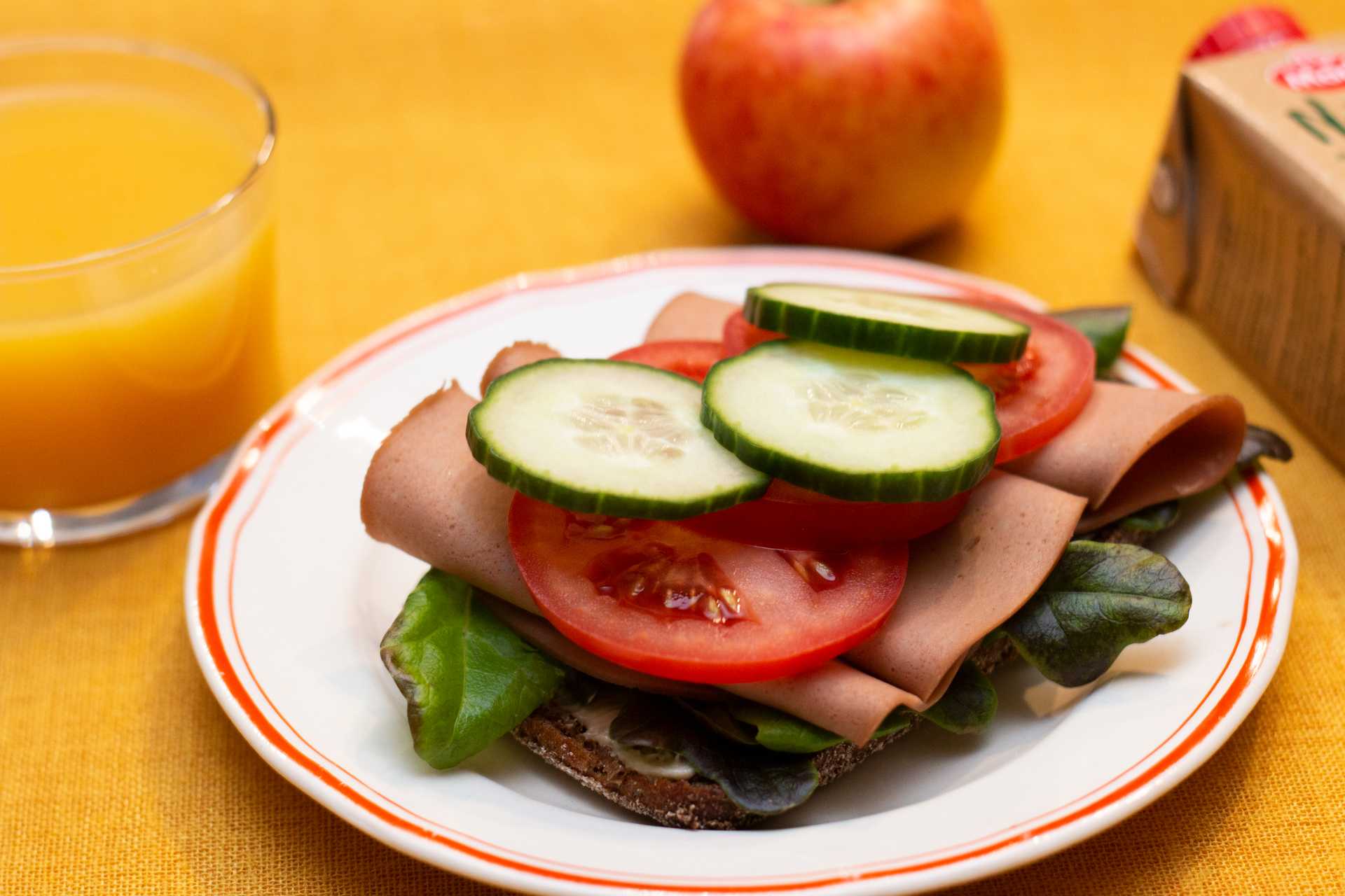 Kuvassa on ruisleipä, jonka päällä on salaattia, vegaanista leikkelettä, tomaattia ja kurkkua. Leivän takana on omena ja vieressä lasillisen appelsiinimehua.