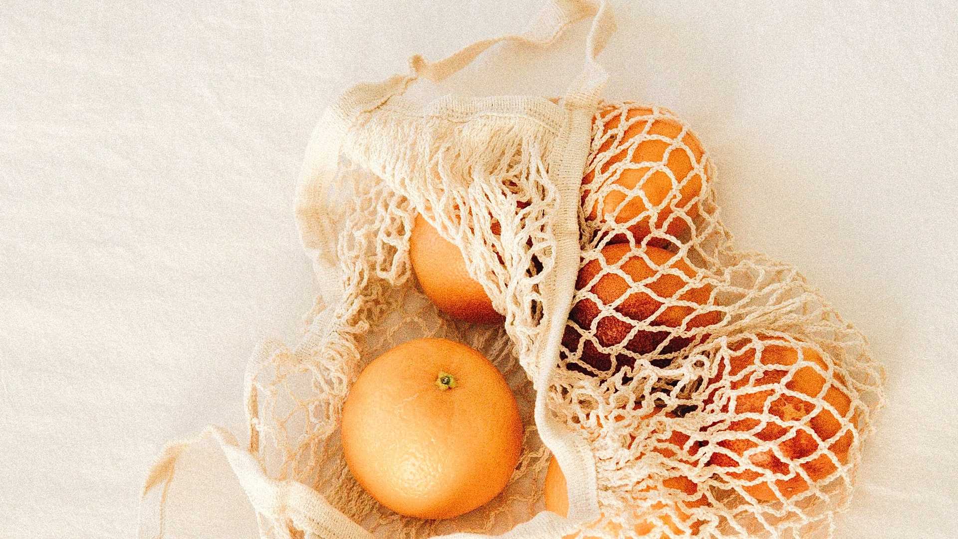 Kuvassa on vaalealla puupöydällä luonnonvalkoinen verkkokassi, jossa on appelsiineja.