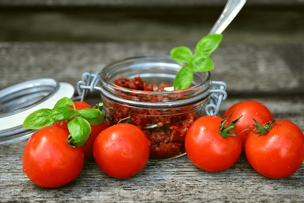 Kuvassa on tuoreita tomaatteja sekä lasikulho, jossa on aurinkokuivattua tomaattia. Tomaaattien päällä on basilikan lehtiä.