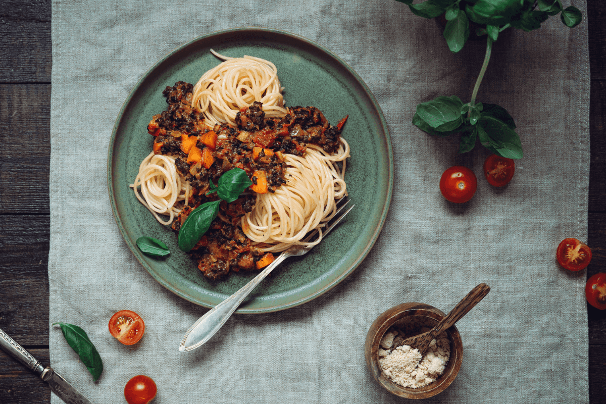 Kuvassa on lautasella spagettia kauniina kiehkuroina sekä soijarouhebolognesekastiketta ja basilikan lehtiä. Lautasen vieressä on puolitettuja kirsikkatomaatteja sekä pieni kulho vegeparmesania.