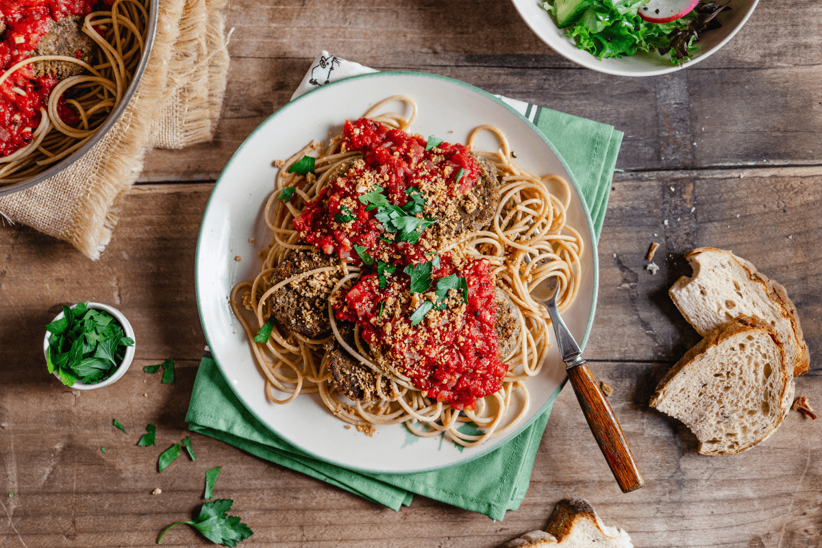 Kuvassa on annos spagettia, tomaattikastiketta ja soijarouhepihvejä. Lautasen vieressä on leipäpaloja, tuoretta lehtipersiljaa sekä salaattiannos.