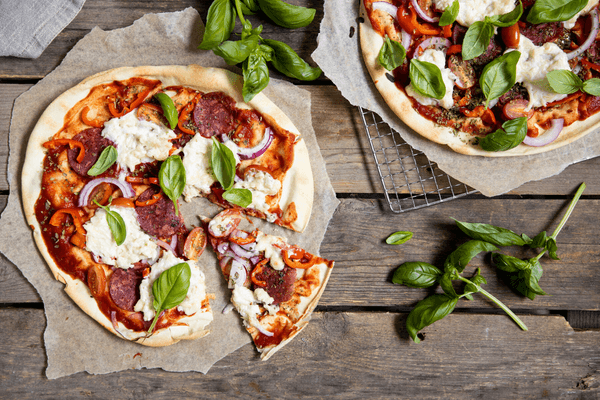 Kuvassa on kaksi pizzaa, joista toinen näkyy vain puolittain. Pizzojen päällä on tomaattikastiketta, vegesalamia, punasipulia, paprikaa ja vegejuustokastiketta. Toisesta pizzasta on leikattu pala. Toinen pizza on ritilän ja toinen revityn leivinpaperin päällä. Niiden vieressä puisella pöydällä on tuoreen basilikan oksia.